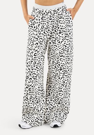 Białe szerokie spodnie z czarnym abstrakcyjnym nadrukiem w panterkę, noszone z białymi trampkami, ręce w kieszeniach, przycięty widok od talii w dół.