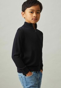 Maglione girocollo blu navy con alto colletto e zip a metà, realizzato in tessuto morbido. Indossato con jeans azzurri chiari, caratterizzati da una vestibilità comoda.