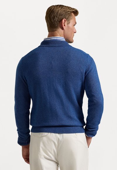 Blå strikket sweater med ribbet krave og manchetter. Har et struktureret mønster, tætsiddende pasform og bæres over lyse bukser.
