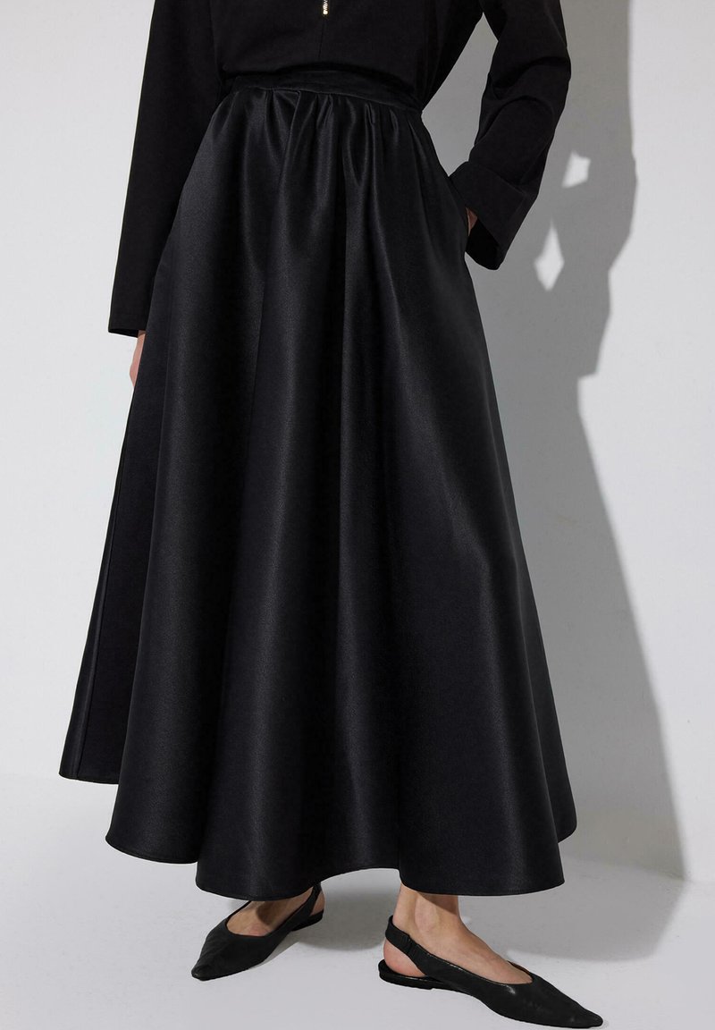 Femme portant une longue jupe noire taille haute plissée, un haut noir et des ballerines noires à bride arrière, debout devant un fond clair uni.