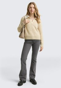 Beżowy sweter polarowy z dekoltem w serek i ściągaczem u dołu, zestawiony z szarymi dzwoniastymi dżinsami i czarnymi butami na platformie. Trzyma beżową torebkę.