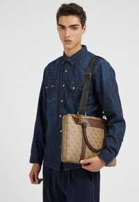 Braune und beige gemusterte Umhängetasche mit einem goldenen Akzent; sie hat eine strukturierte Form und einen verstellbaren Tragegurt und wird von einer Person in einem Jeanshemd gehalten.