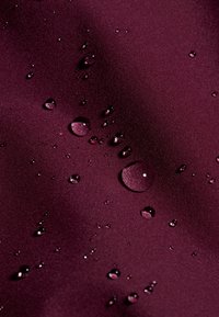 Tessuto bordeaux con una texture liscia, caratterizzato da varie gocce d'acqua di dimensioni diverse che riflettono la luce sulla superficie.