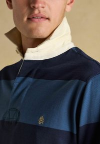 Nahaufnahme einer Person, die ein navyblaues und blau gestreiftes Rugby-Shirt mit einem weißen hochgeklappten Kragen und einem kleinen gestickten goldenen Logo auf der Brust trägt.