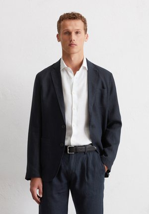 Marineblaues Leinen-Blazer mit zwei Knöpfen, passendem weißen Leinenhemd, schwarzem Leder-Gürtel und dunklen Hosen. Verfügt über eine Vordertasche am Blazer.