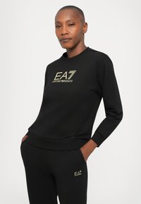 Rövid hajú személy fekete EA7 Emporio Armani pulóverben és azzal megegyező nadrágban, zsebekbe tett kézzel, egyszínű háttér előtt.