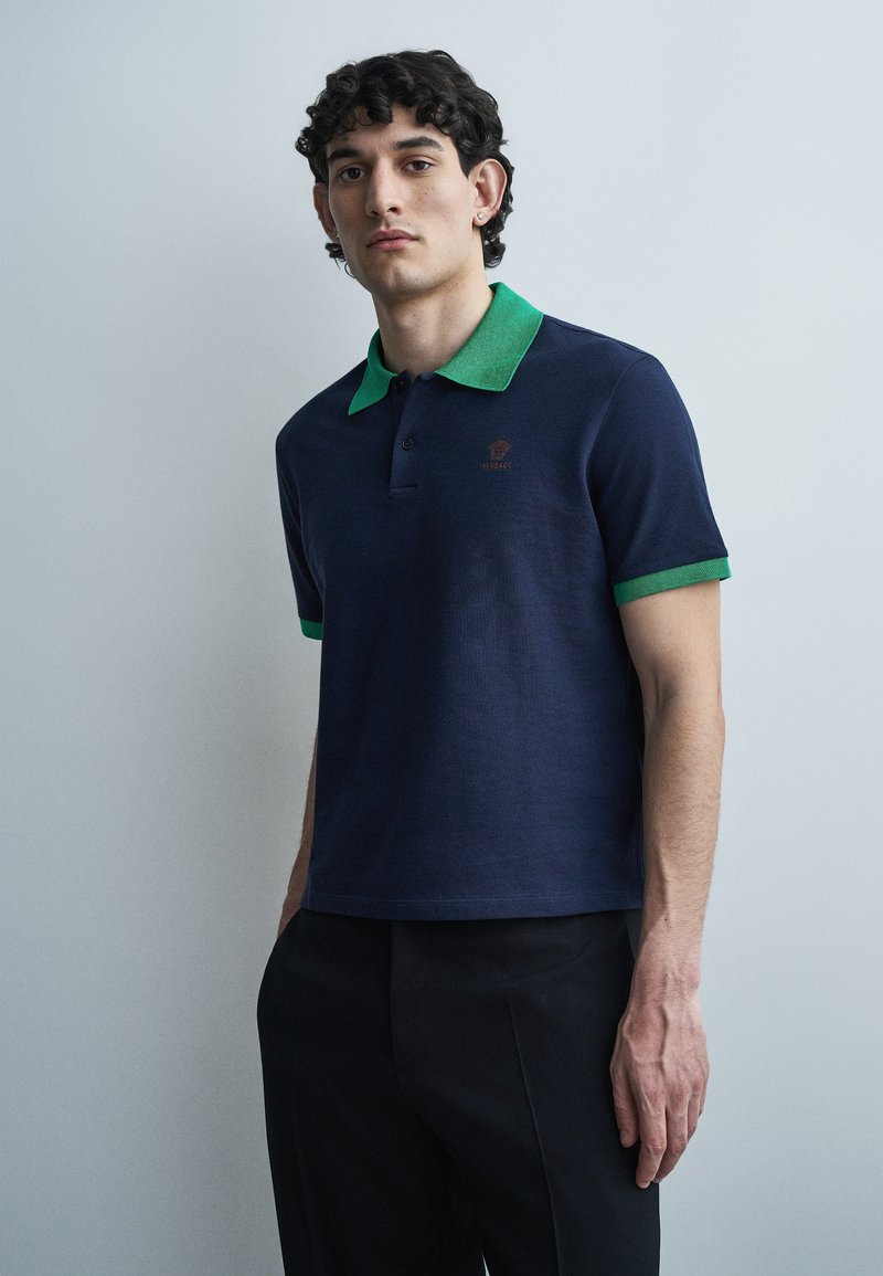 Jonge man met een donkerblauw poloshirt met groene kraag en mouwranden, staande tegen een lichtgrijze achtergrond.