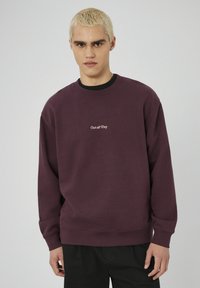 Jeune personne avec des cheveux blonds courts portant un sweat à capuche violet foncé avec le texte "Sorti toute la journée", se tenant devant un fond clair.