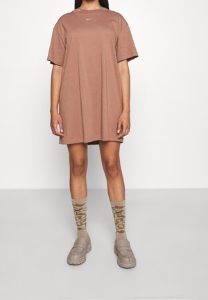 Kobieta ubrana w oversize'ową brązową sukienkę t-shirt Nike, beżowe wzorzyste skarpetki Nike oraz beżowe masywne loafersy, stojąca na białym tle.