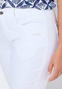 Los jeans de mezclilla blanca presentan una textura suave, un ajuste slim y un sutil bordado de "lovel" en el costado, combinados con una blusa estampada.