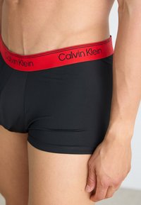 Zwarte katoenen boxershorts met een rode elastische tailleband met het logo "Calvin Klein". Close-up van de stofstructuur en pasvormdetails.