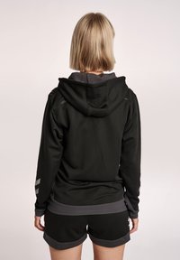 Felpa nera con fodera interna grigia e dettagli in mesh, dotata di cappuccio e strisce riflettenti sulle maniche. Abbinata a pantaloni corti neri.
