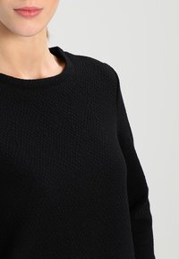Sort struktureret sweater med rund halsudskæring og lange ærmer, der har et vævet mønster for ekstra dybde og visuelt fokus.