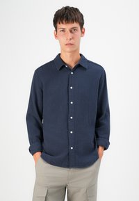 Navyblauwe longsleeve blouse met een gestructureerd patroon, knoopsluiting en een relaxte kraag. Voorzien van witte knopen en opgerolde mouwen.