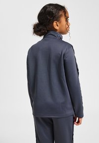 Graue Sportjacke mit hohem Kragen, glatter Textur und kontrastierenden Seitenstreifen. Hat eine lockere Passform und lange Ärmel.