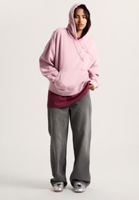 Oversized pink hoodie with a front pocket, featuring a drawstring hood. Worn over a burgundy shirt and paired with wide gray jeans.