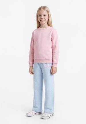 Ragazza giovane con lunghi capelli biondi che indossa una felpa rosa, pantaloni azzurri con testo "Amour" e scarpe da ginnastica bianche, in piedi e sorridente.