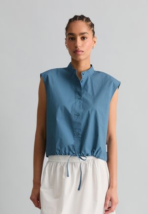 Kobieta z warkoczami, ubrana w niebieski, bezrękawnikowy top zapinany na guziki z wiązaniem w talii oraz białą spódnicę, zwrócona przodem na jednolitym tle.