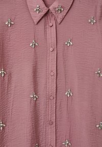 Camicia rosa con texture e colletto, caratterizzata da decorazioni argentate che riproducono fiori, e una fila di bottoni sul davanti.