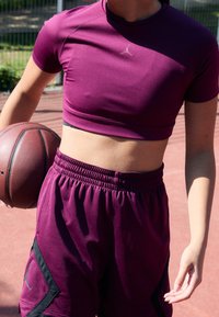 Osoba v purpurovém sportovním oblečení držící basketbal, stojící na venkovním basketbalovém hřišti ve dne.