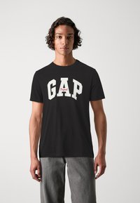 Camiseta negra con el logo "GAP" en blanco y acento rojo, de manga corta y cuello redondo; la suave tela de algodón brinda comodidad.