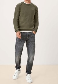 Khaki Rundhalspullover mit gerippten Bündchen, kombiniert mit ausgewaschenen blauen Jeans. Das Modell trägt weiße Sneaker. Einfacher und entspannter Freizeitlook.