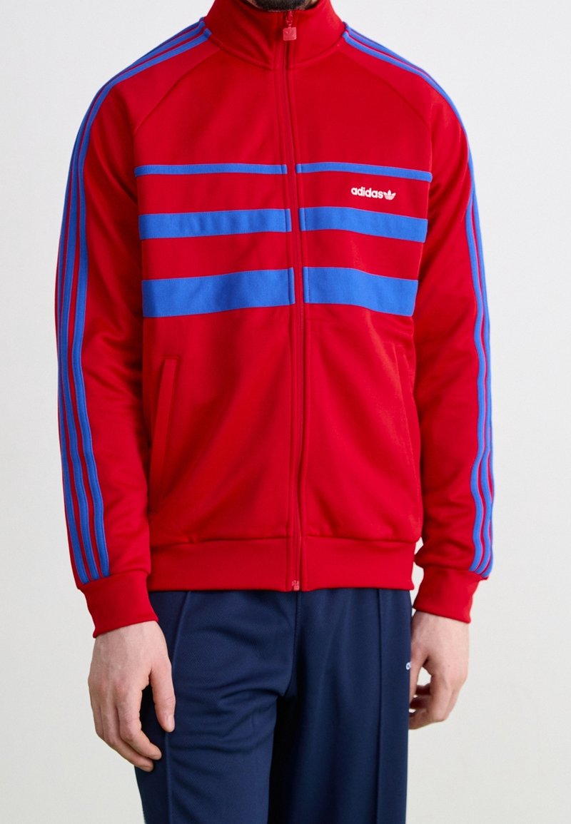 Červená sportovní bunda na zip s modrými vodorovnými pruhy, s límcem do stoje a dvěma bočními kapsami; vyrobená z hladkého sportovního materiálu.