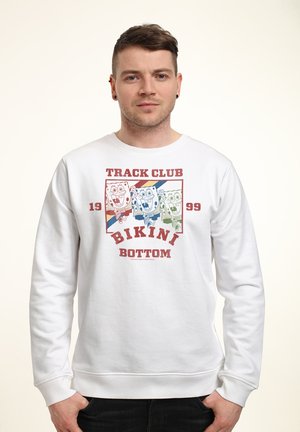 Giovane uomo che indossa una felpa bianca con grafica colorata di SpongeBob SquarePants che corre e testo "Track Club Bikini Bottom 1999."