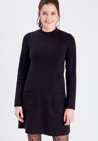 Robe noire à manches longues avec un col rond, dotée de deux poches avant et d'une coupe droite. Texture en tricot lisse et longueur genou.