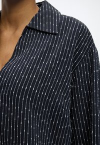 Gros plan d'une personne portant une chemise bleu foncé avec des rayures verticales blanches ponctuées et un col ouvert sur un fond uni.