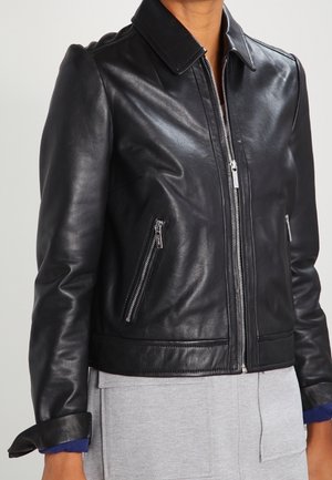 Veste en cuir noire avec fermeture éclair devant et poches latérales zippées portée sur un vêtement gris.