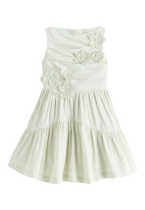 Junges Mädchen mit einem Seitenzopf, das ein helles ärmelloses Kleid mit floralen Details und passende Sandalen trägt, läuft vor einem einfarbigen Hintergrund.
