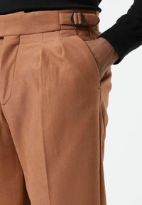 Bruine op maat gemaakte broek met een gladde textuur, plooien en een lussensluiting met een zwarte metalen gesp. Zijkantzak zichtbaar.