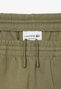 Pantalones de pullover en verde oliva con cintura elástica, cordón y una etiqueta que muestra "Lacoste Regular Fit". Etiqueta con detalles legibles a máquina visible.