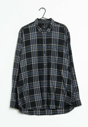 Camisa de cuadros larga de manga larga en negro, azul y blanco, colgada en una percha negra contra un fondo blanco.