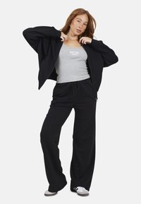 Haut à zip noir sur un débardeur gris avec le texte "NEW YORK", associé à un pantalon noir à cordon de serrage et à des baskets grises. Tissu lisse, coupe décontractée.