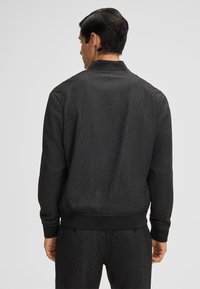 Giacca bomber nera con una leggera lucentezza, polsini e orlo elastici, texture liscia, design a coda di giacca e un piccolo dettaglio con bottone sul collo.