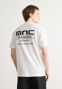 Hvit bomull t-skjorte med korte ermer, med svart trykt tekst på baksiden: "mnc," "Mads Nørgaard," "COPENHAGEN," og kontaktinformasjon.