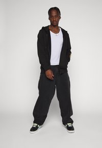 Sudadera negra con cremallera sobre una camiseta blanca, combinada con jeans negros holgados y zapatillas negras con detalles verdes, de pie contra un fondo gris.