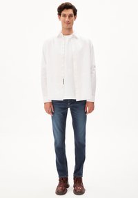 Camisa blanca de botones sobre una camiseta blanca, combinada con jeans azules y zapatillas marrones. El conjunto presenta un ajuste informal y líneas limpias.