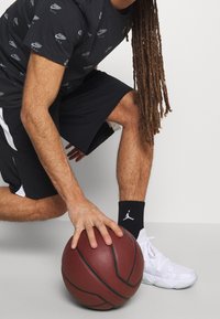 Černé sportovní tričko s opakujícím se vzorem loga, černé kraťasy, bílé tenisky a černé ponožky s logem, které drží basketbal v dřepu.