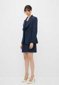 Marineblauer maßgeschneiderter Blazer mit breitem Revers, zwei Knöpfen und Fronttaschen, kombiniert mit einem passenden Rock. Glattes Material und strukturiertes Design.