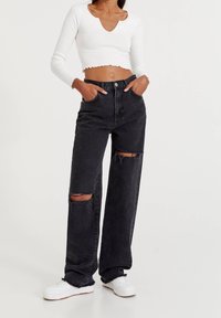 Jeans en denim noir taille haute à jambes larges, avec des déchirures aux genoux et des ourlets bruts. Associés à un haut blanc côtelé court.