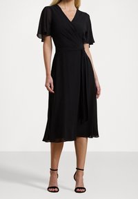 Svart, knälång omslagsklänning med korta volangärmar, V-ringning och midjeband. Tillverkad av lätt, mjukt tyg med en slät textur.