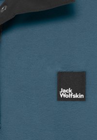 Giacca in fleece di colore blu, con una texture morbida. Etichetta nera con il testo bianco "Jack Wolfskin" cucita nell'angolo in basso a destra.