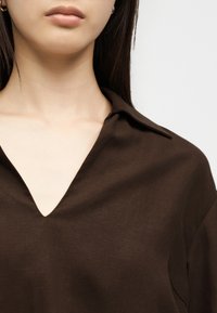 Κοντινό πλάνο ενός ατόμου που φοράει ένα σκούρο καφέ T-shirt με V-λαιμόκοψη και γιακά, εμφανίζοντας το λαιμό, το πηγούνι και μέρος των μακριών σκούρων μαλλιών.