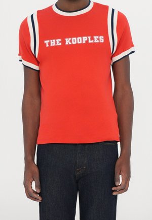 Mężczyzna w czerwonej koszulce z krótkim rękawem z białymi i niebieskimi paskami na rękawach oraz napisem "THE KOOPLES" na piersi, w ciemnych dżinsach.