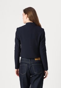 Blazer cropped navy con spalle strutturate, linee pulite e colletto a punte. Indossato su jeans scuri per creare contrasto.