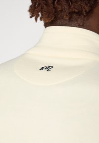 Lys cremefarvet pullover med en blød tekstur, der har en høj krave og et sort broderet logo på området bagpå halsen.