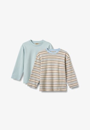Rødharret barn smilende, iført en lyseblå langærmet skjorte, blå jeans og beige slip-on sko, står mod en hvid baggrund.